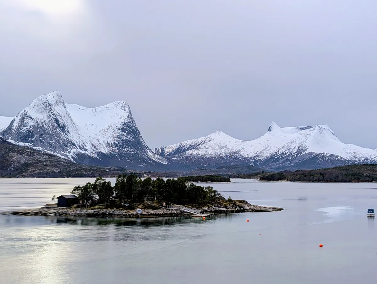 Pause photo sur la E6 entre Narvik et Bodø : un petit îlot boisé avec des cabanes, encadré par les sommets enneigés du Nordland.