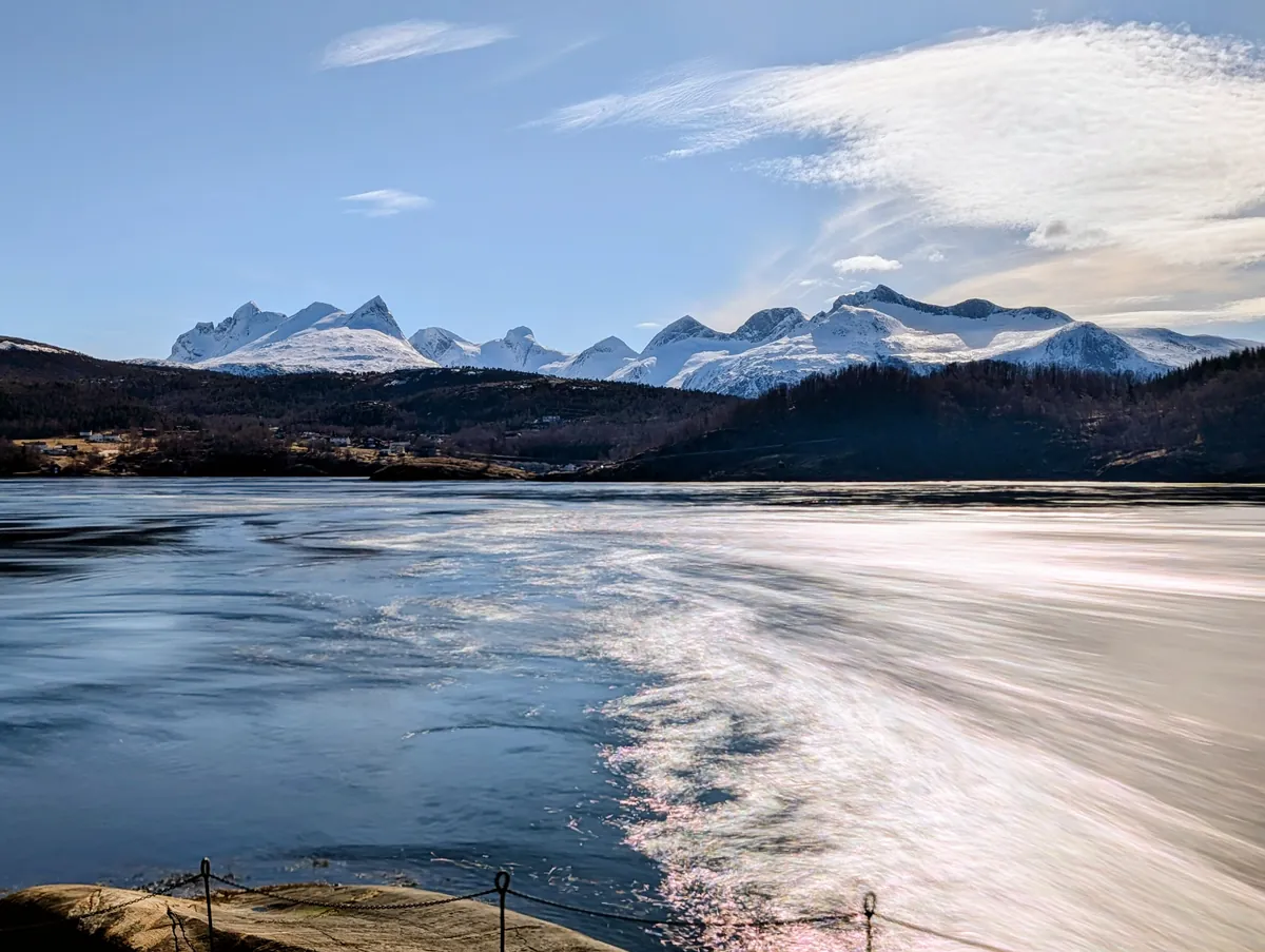 Vue large sur le courant du Saltstraumen en pose longue depuis la rive — l'eau qui « bout » avec les montagnes du Børvasstindan en arrière-plan.
