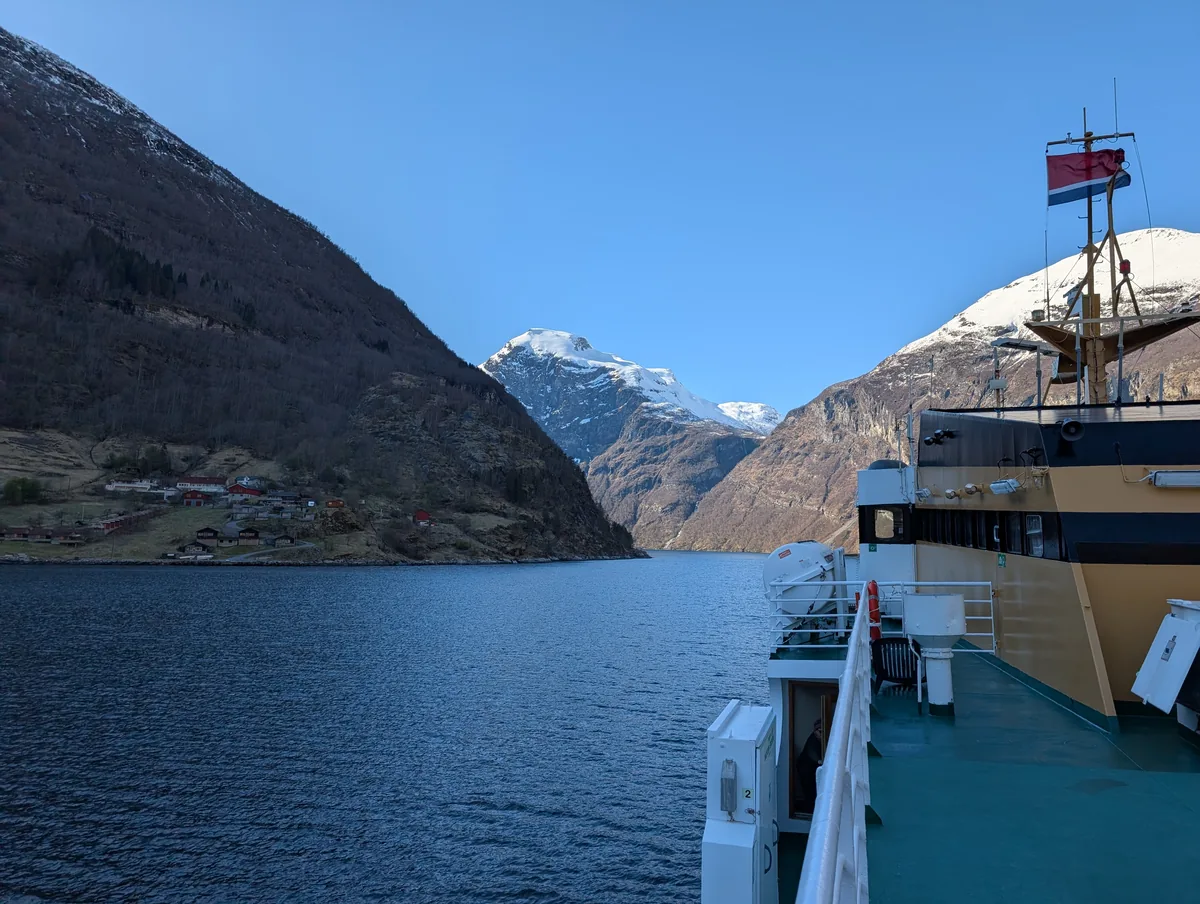 Drapeau norvégien claquant au vent à l'arrière du ferry, avec le Geirangerfjord et ses falaises enneigées en toile de fond.