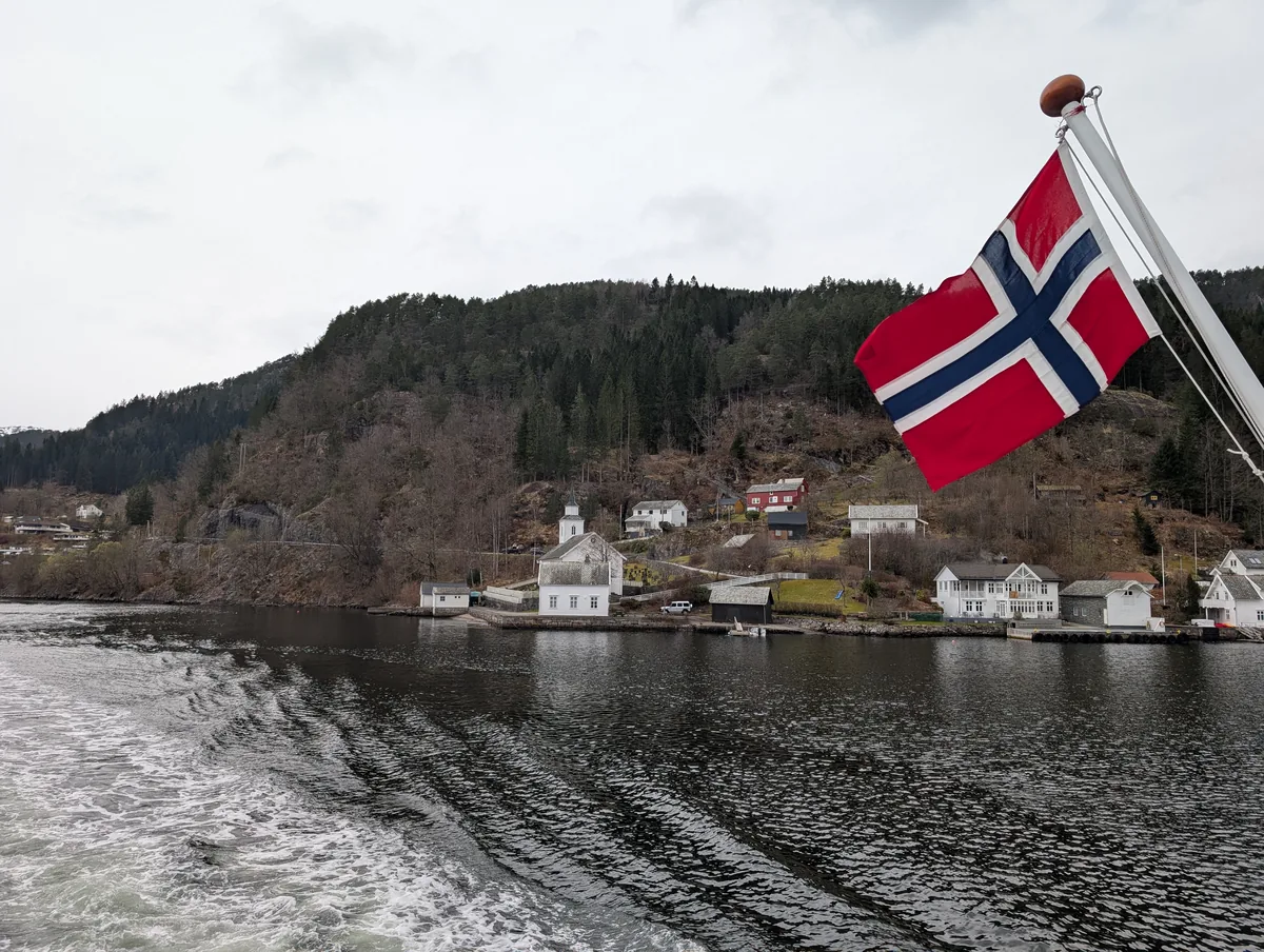 Maisons traditionnelles sur les rives du fjord Petites maisons traditionnelles norvégiennes sur les rives boisées de l'Osterfjord, drapeau norvégien au vent à l'arrière du bateau de croisière.