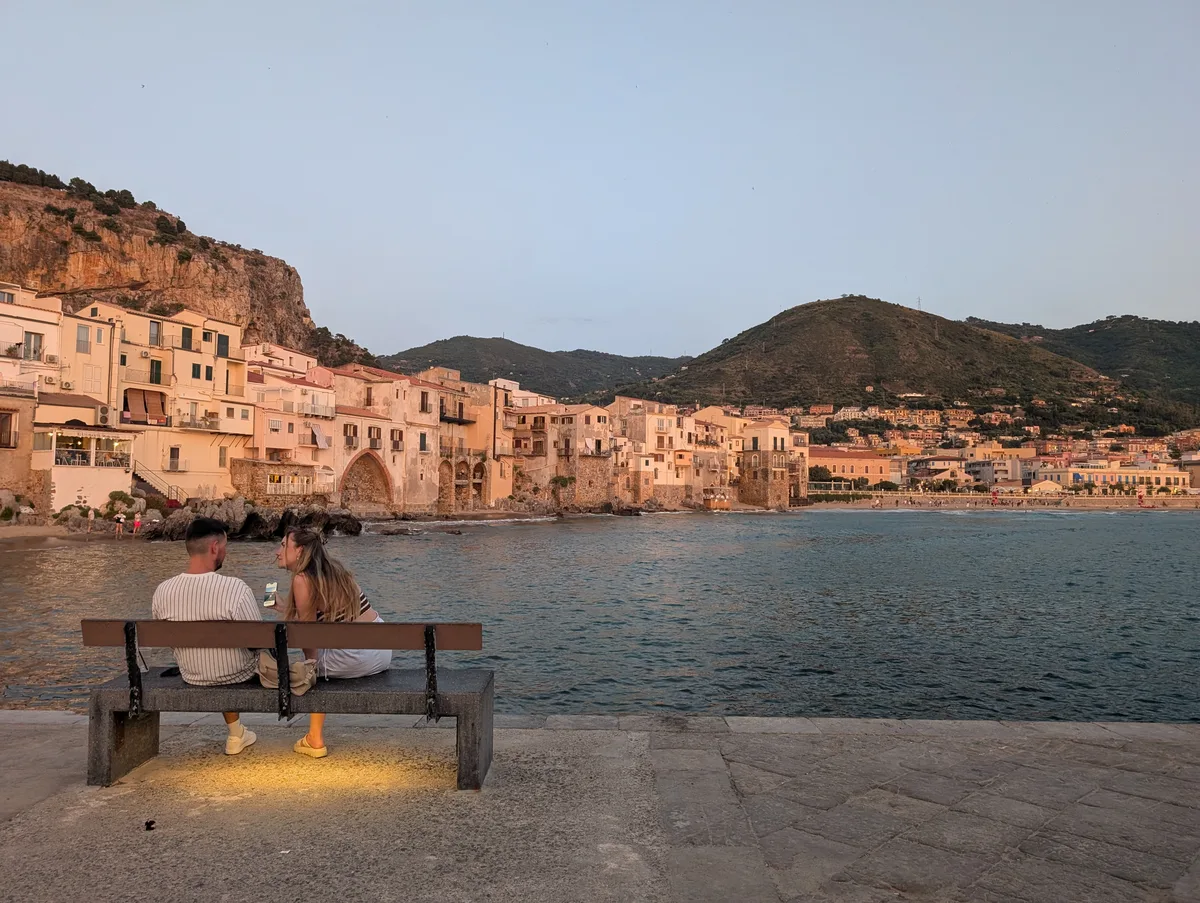 Un couple assis sur un banc face à la mer contemple le coucher de soleil sur la vieille ville de Cefalù en Sicile.