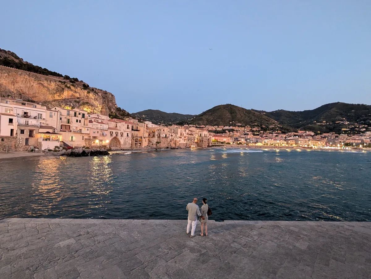 Vista nocturna del paseo marítimo de Cefalù con una pareja de pie a orillas del mar Vista nocturna del paseo marítimo de Cefalù con una pareja de pie a orillas del mar