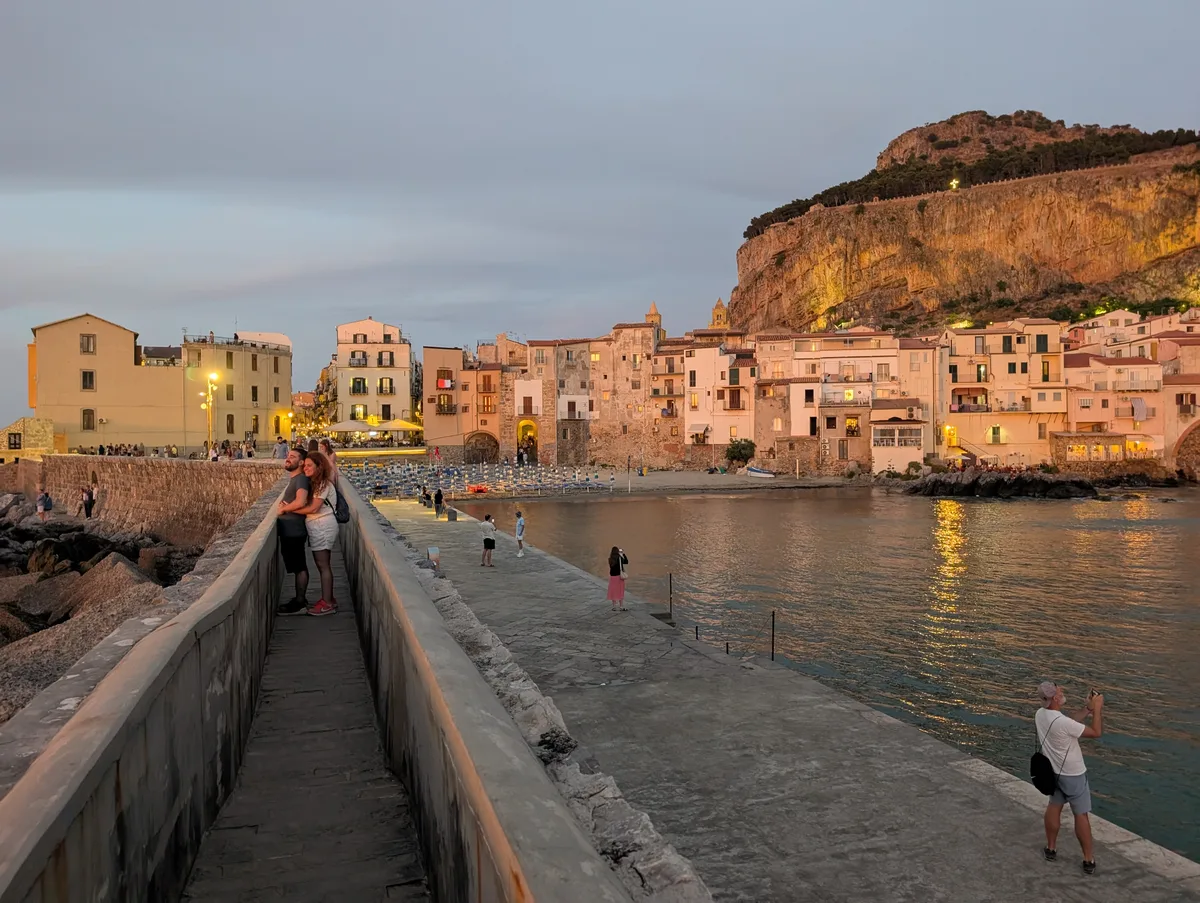 Paseantes en el espigón de Cefalù al anochecer con el casco antiguo y la Rocca Paseantes en el espigón de Cefalù al anochecer con el casco antiguo y la Rocca