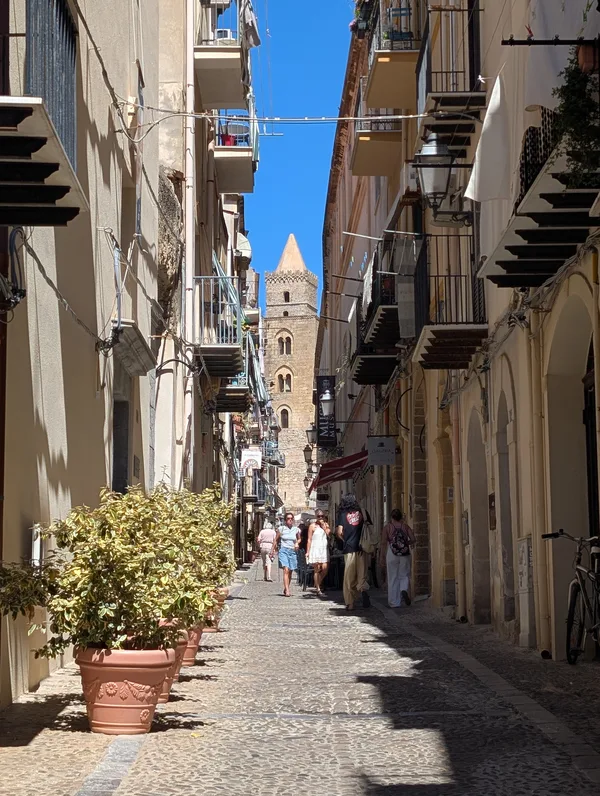 Ruelle typique du centre historique de Cefalù avec des pots de fleurs et la cathédrale normande visible en arrière-plan.