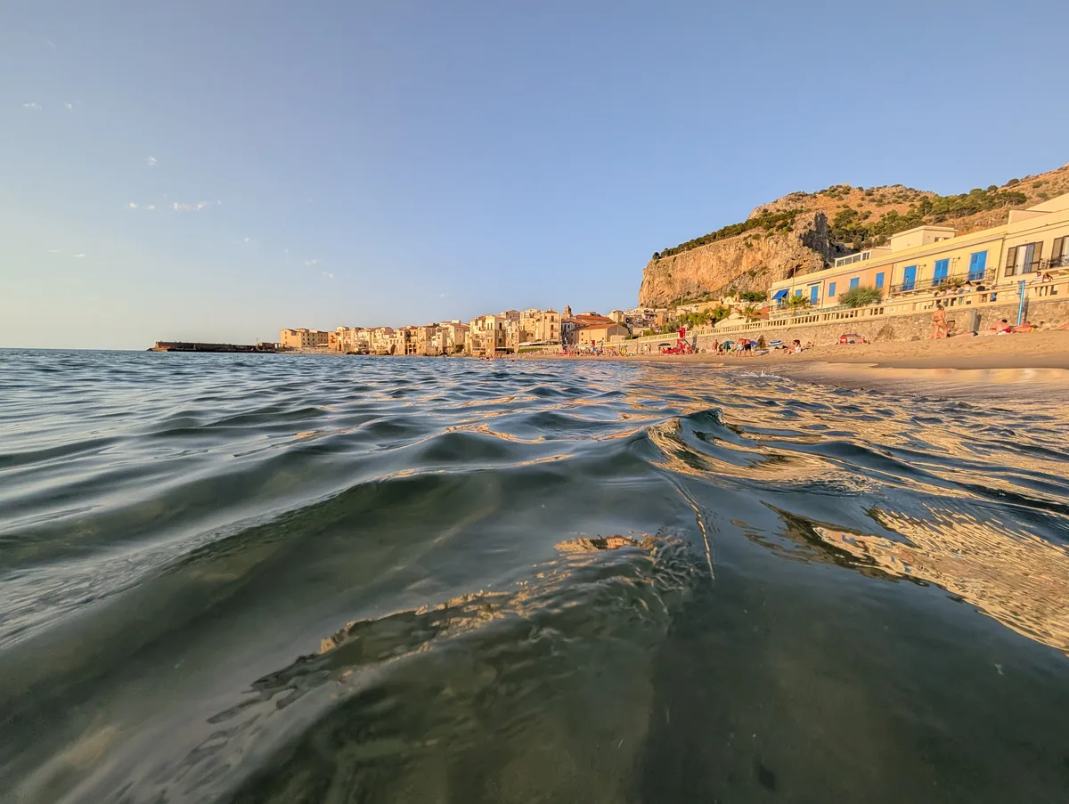 Vue mer sur Cefalù et la Rocca au coucher de soleil Vue depuis la mer sur la vieille ville de Cefalù et la Rocca au coucher de soleil, avec des vagues au premier plan en Sicile.