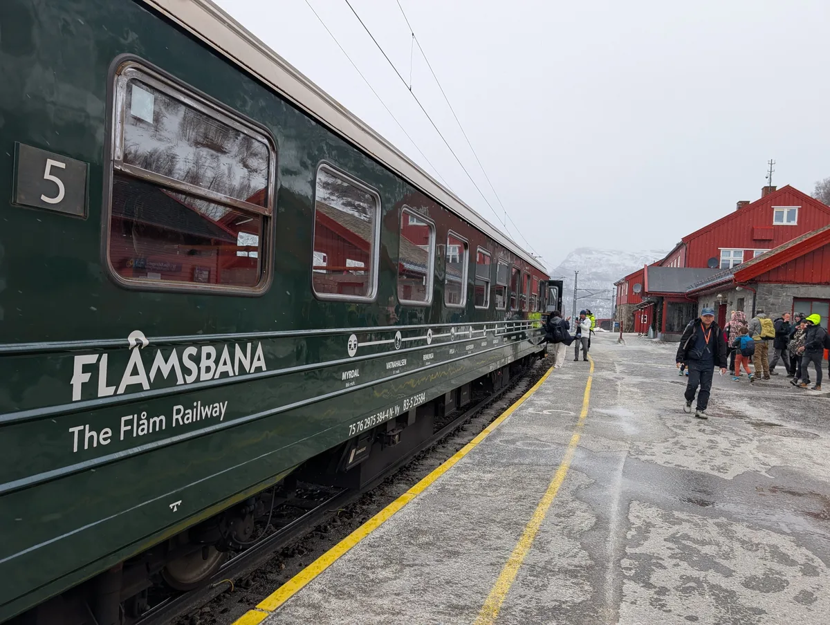 Wagon vert numéro 5 du Flåmsbana à quai en gare de Myrdal sous la neige, voyageurs marchant devant les bâtiments rouges.