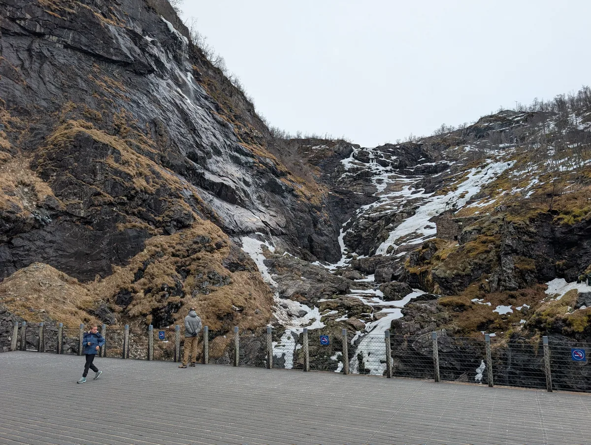 Voyageur sur la plateforme d'observation de Kjosfossen face à la paroi rocheuse enneigée, lors de l'arrêt du train de Flåm.