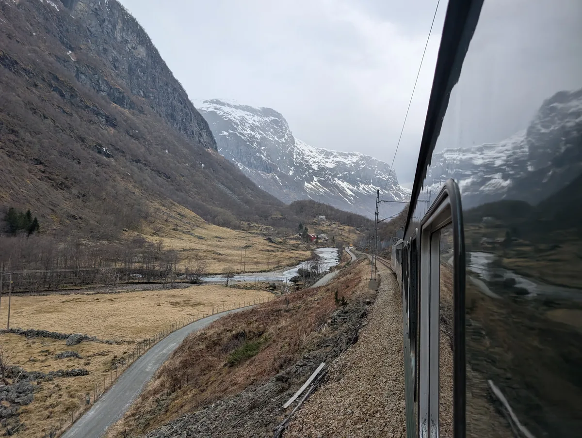 Le train de Flåm longe la rivière au fond de la vallée, ses wagons verts dessinant une courbe sous les montagnes enneigées.