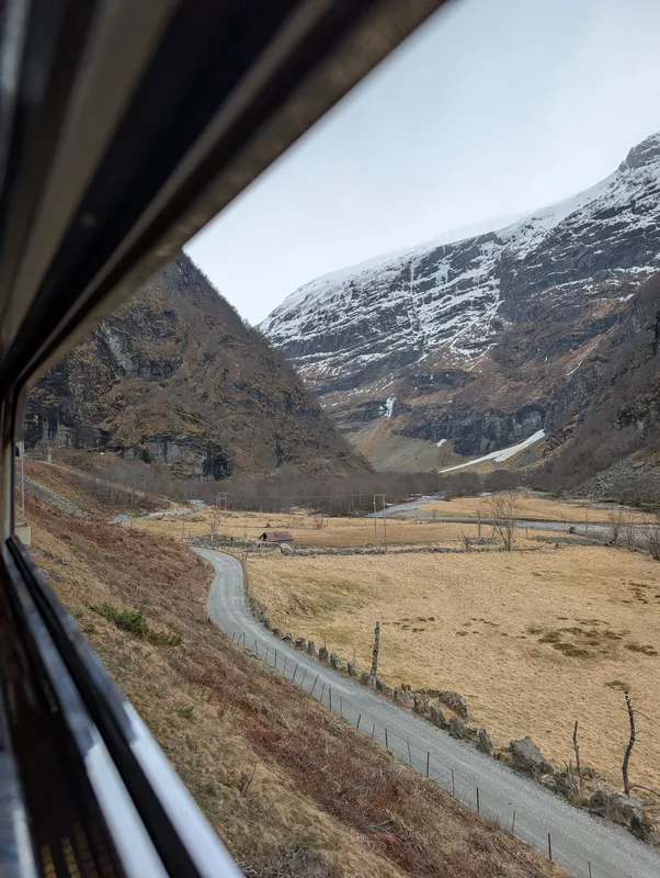 Vue plongeante depuis la fenêtre du train de Flåm sur une route sinueuse traversant la vallée vers les montagnes enneigées.