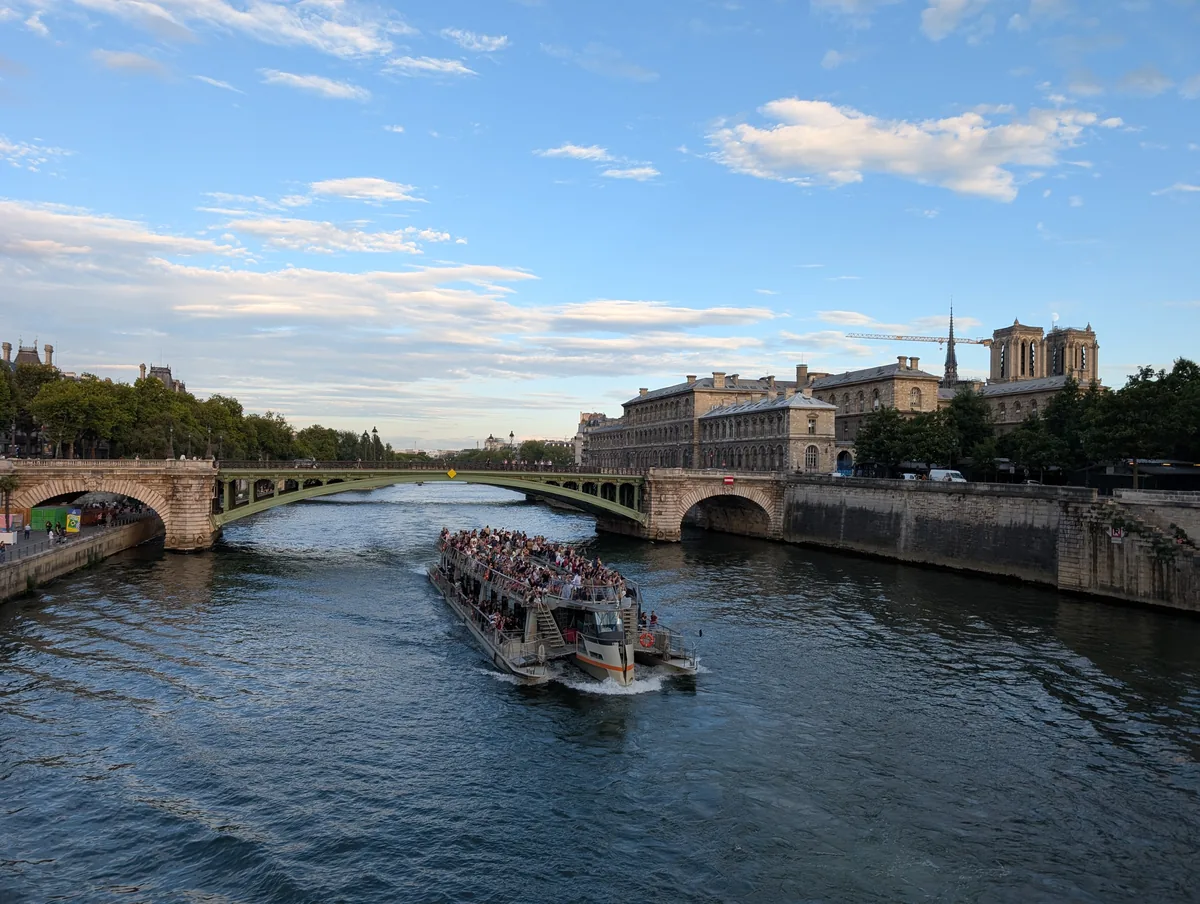 Bateau de croisière chargé de passagers franchissant le pont Notre-Dame sur la Seine, cathédrale visible en arrière-plan à Paris.