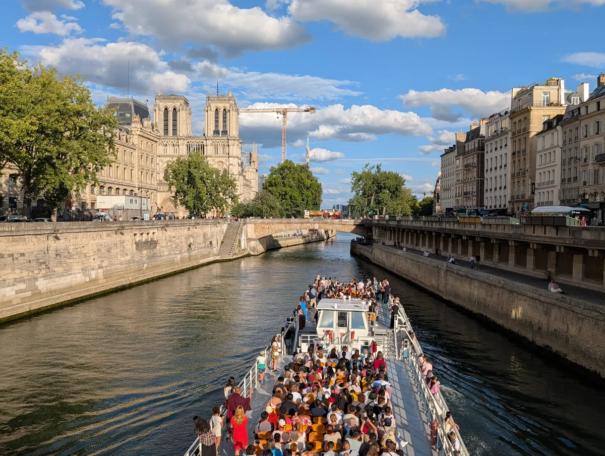 Pont supérieur d'un bateau de croisière rempli de passagers passant devant la cathédrale Notre-Dame de Paris, par ciel bleu.
