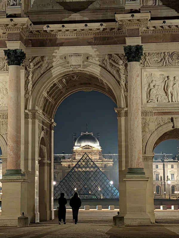 The Carrousel arch and the Louvre pyramid by night The Louvre pyramid framed by the Arc de Triomphe du Carrousel at night, with two strolling figures in Paris.