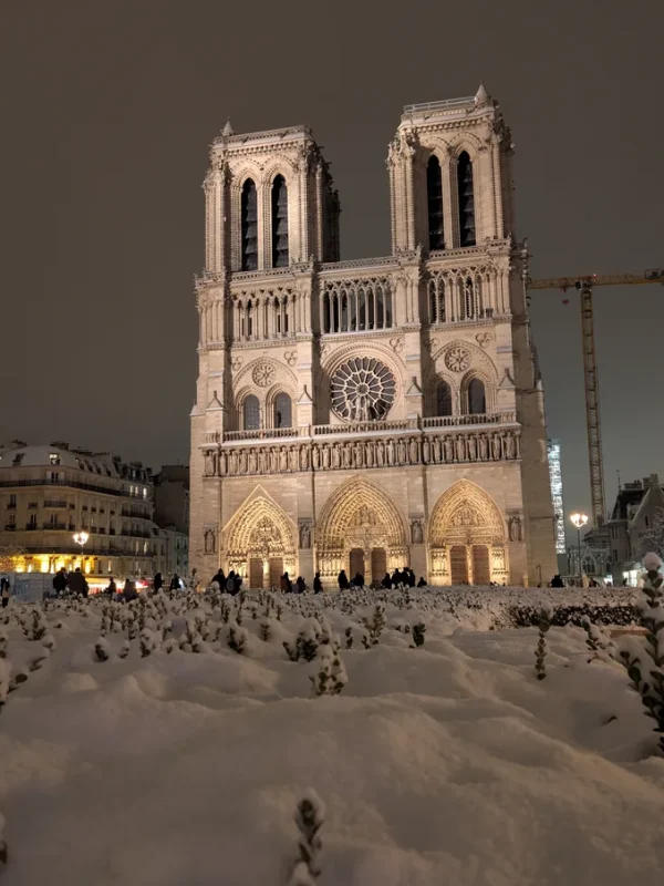 Notre-Dame de Paris under the snow at night The floodlit façade of Notre-Dame cathedral in Paris at night, seen from a square entirely covered in snow.