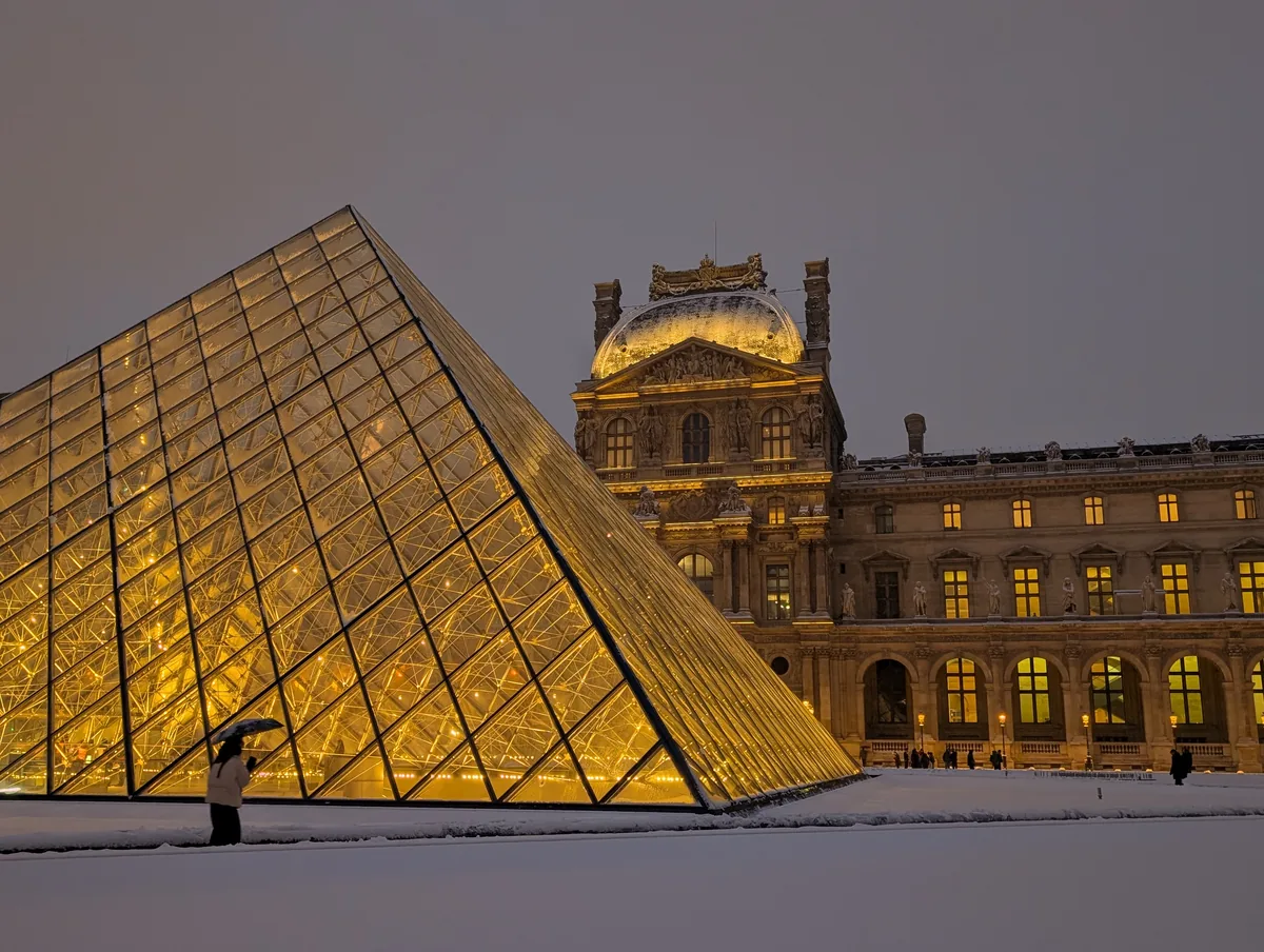 The Louvre pyramid under the snow in Paris The Louvre pyramid lit up at dusk in a Cour Napoléon entirely covered in snow, a passer-by in the foreground in Paris.