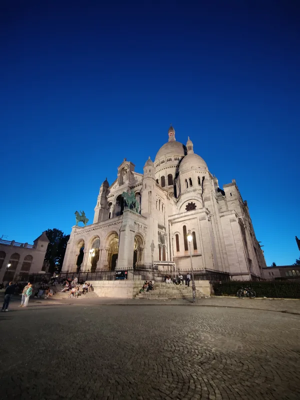 The Sacré-Cœur basilica at blue hour The floodlit façade of the Sacré-Cœur basilica in Montmartre at blue hour, with visitors sitting on the steps in Paris.