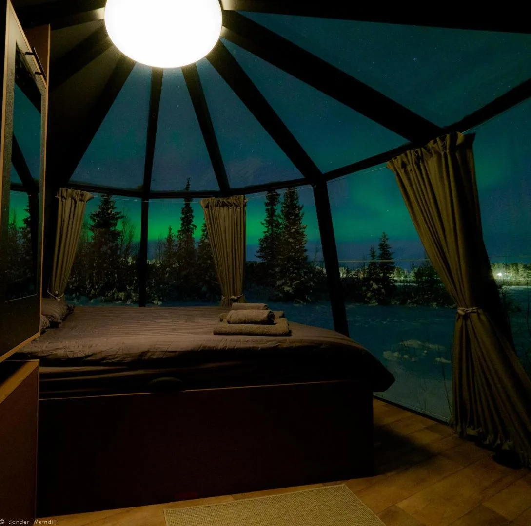 Cama de un iglú de cristal del Aurora River Camp cerca de Kiruna, con una aurora boreal verde sobre el bosque nevado