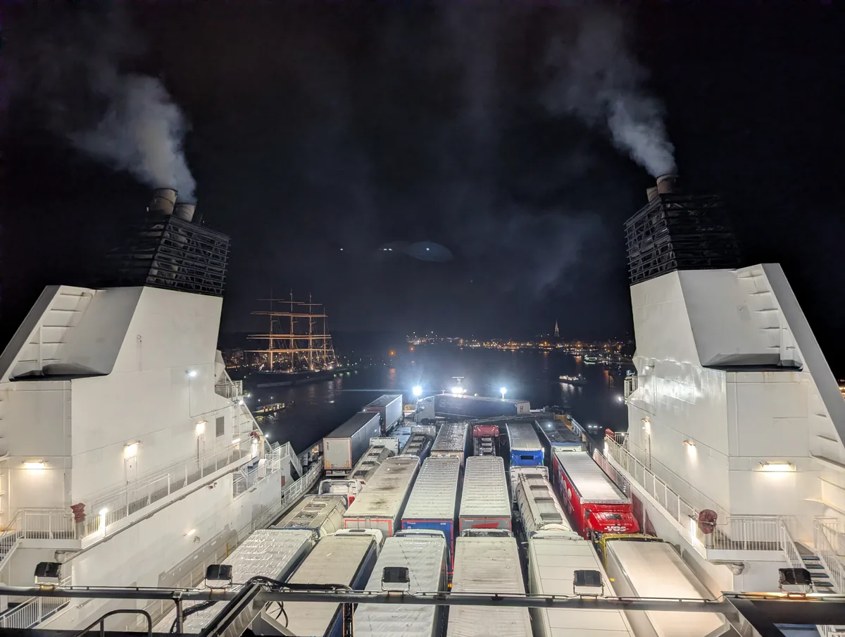Rauchende Schornsteine der Fähre Tinker Bell und Lkw auf dem Achterdeck, mit der Passat und Travemünde in der Ferne bei der Abfahrt