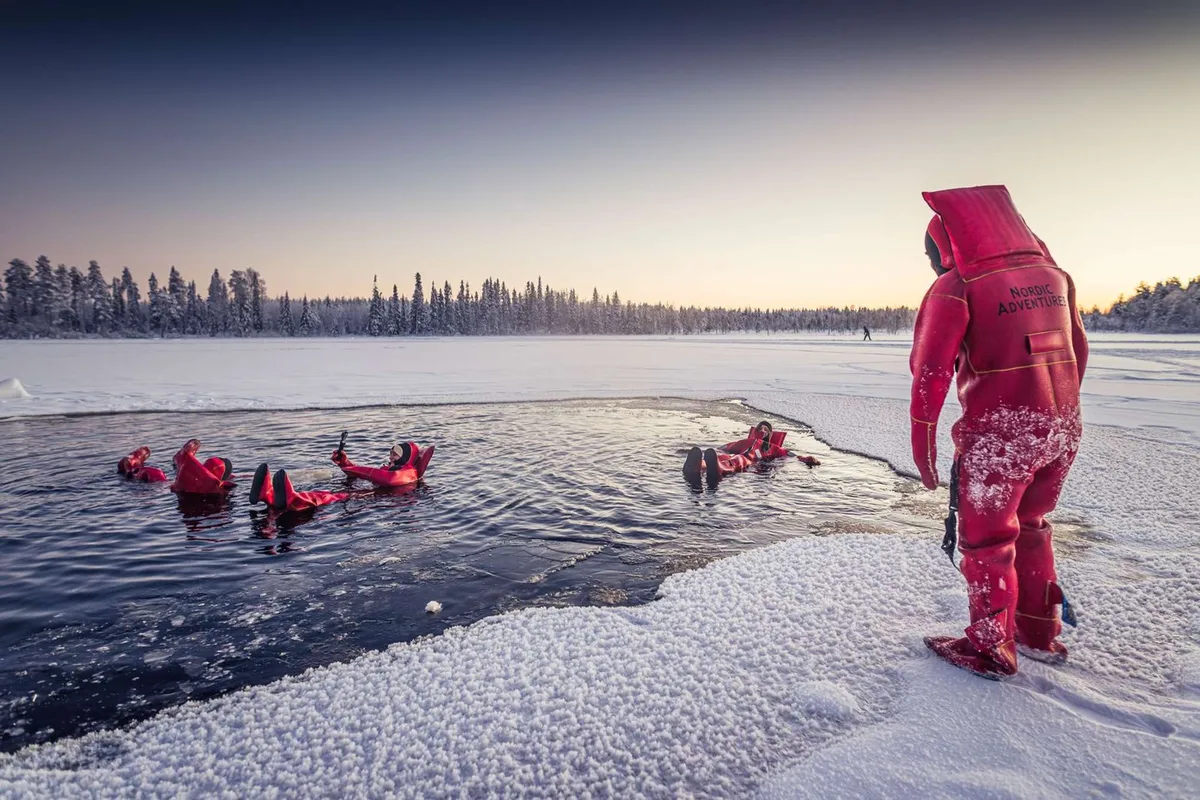 Ice floating on a frozen lake near Rovaniemi with Nordic Adventures: people in survival suits floating on their backs in the icy Lapland water surrounded by snow, heated cabin on the way back
